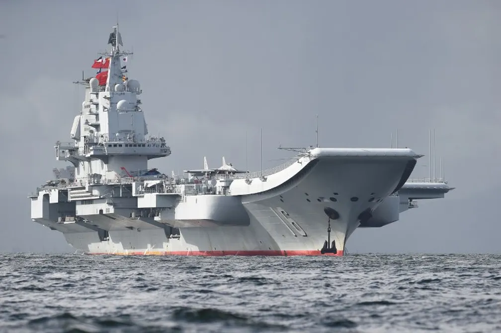 Navio militar chinês patrulhando águas disputadas no Mar do Sul da China, com o céu ao fundo e a presença de infraestrutura marítima próxima.
