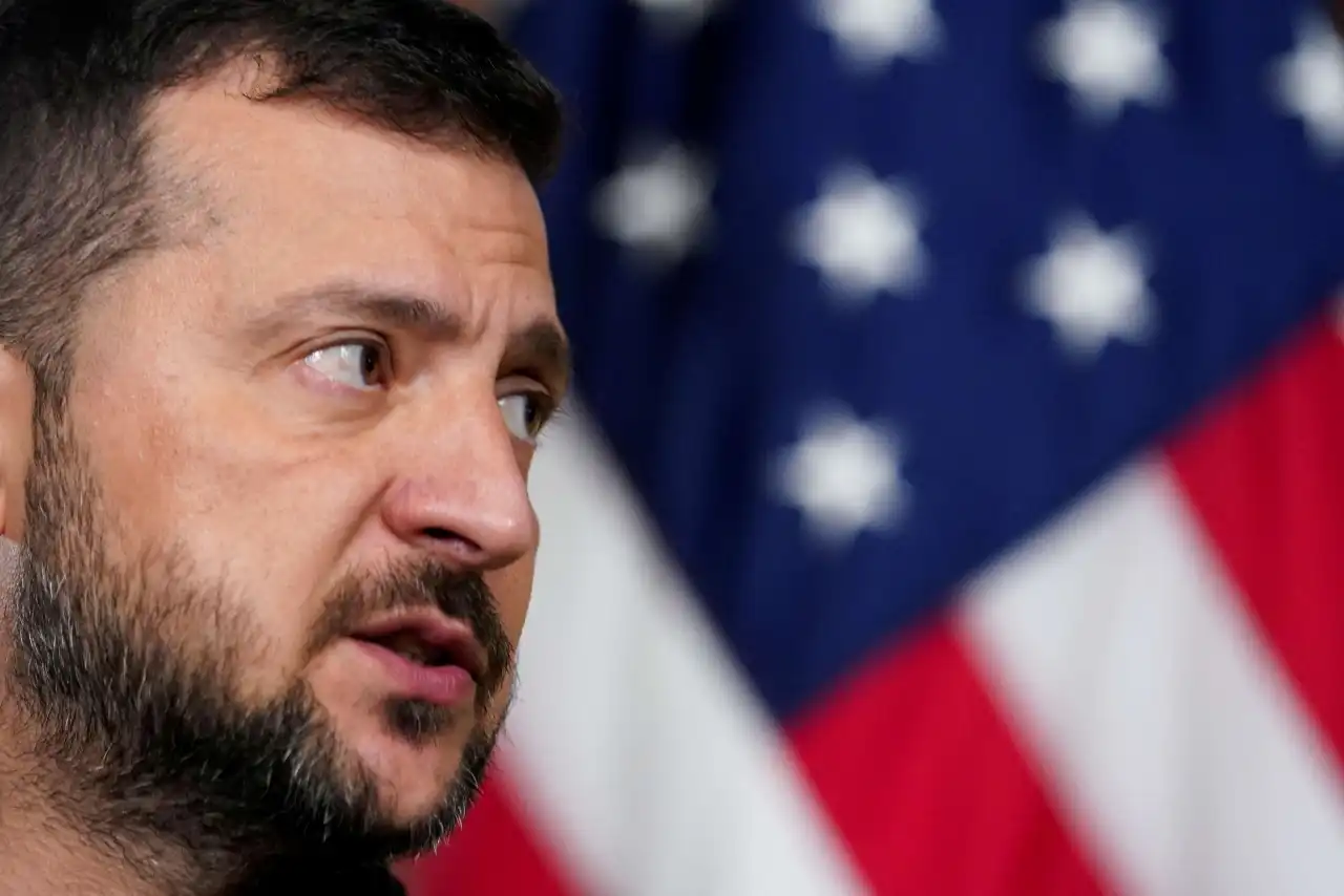 Imagem de Volodymyr Zelensky durante uma coletiva de imprensa, com expressão séria, comentando sobre o papel dos Estados Unidos no conflito na Ucrânia.