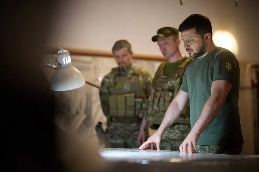 Zelensky em uma sala de reuniões com dois oficiais militares, observando atentamente um mapa, enquanto discutem a proposta de envio de tropas ocidentais para a Ucrânia.