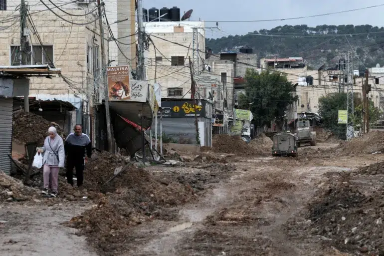 Palestinos caminham por uma estrada danificada perto da entrada principal do campo de refugiados de Jenin durante a operação militar israelense em andamento na área norte da Cisjordânia ocupada, 5 de fevereiro de 2025 [Alaa Badarneh/EPA] Fonte: Al Jazeera