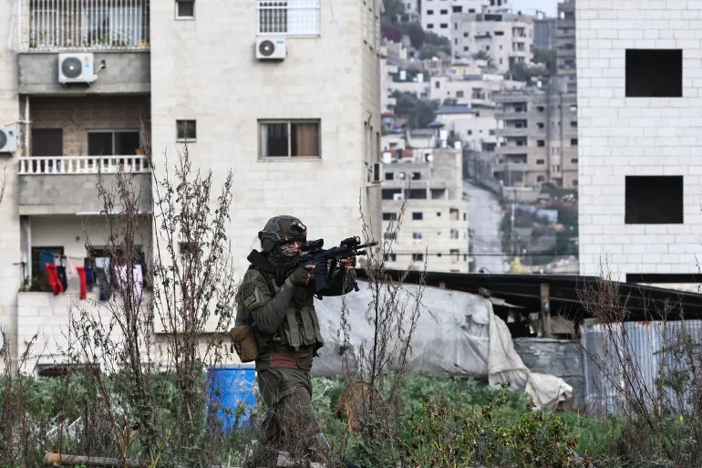 Um soldado israelense aponta seu fuzil enquanto está em um campo na entrada do campo de refugiados de Tulkarem, 5 de fevereiro de 2025 [Zain Jaafar/AFP] Fonte: Al Jazeera