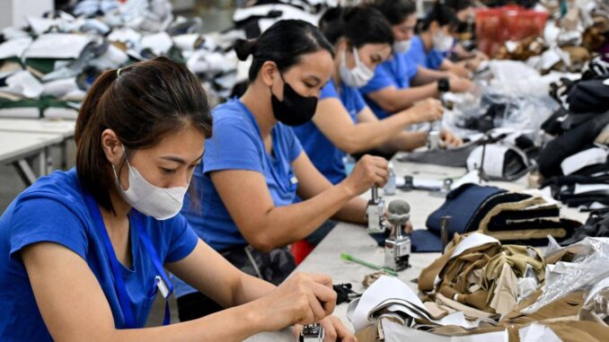 2025-07-10-vietnam-factory-tariff-2222465468 Trabalhadoras vietnamitas em fábrica têxtil usando máscaras, costurando peças de roupa.
