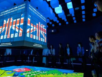 Visitantes observando uma instalação interativa tecnológica com projeções digitais coloridas em uma sala escura.