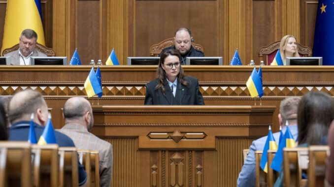 2025-07-22-ukraine-parliament-Svyrydenko-2224813694 Yulia Svyrydenko discursando no Parlamento ucraniano durante a sessão de reformulação do governo, com bandeiras da Ucrânia e da União Europeia ao fundo.