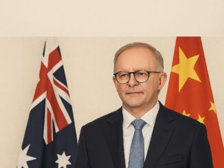 Primeiro-Ministro australiano Anthony Albanese em pé, vestindo terno escuro, gravata azul e óculos, com as bandeiras da Austrália e da China ao fundo.