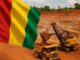Bandeira da Guiné tremulando em primeiro plano, com uma grande escavadeira carregando bauxita em uma mina de terra vermelha ao fundo.
