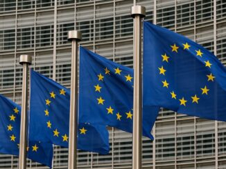 Bandeiras da União Europeia tremulando ao vento em frente a um edifício moderno com janelas de vidro.