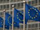 Bandeiras da União Europeia tremulando ao vento em frente a um edifício moderno com janelas de vidro.
