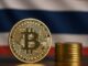 Moeda física dourada de Bitcoin em destaque sobre uma superfície de madeira, com uma pilha de moedas ao fundo e uma bandeira desfocada com cores vermelho, branco e azul.