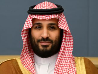 Retrato de um homem árabe usando traje tradicional saudita com keffiyeh vermelha e branca e thobe branco, sorrindo levemente.