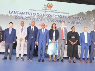 Grupo de doze pessoas, entre homens e mulheres, em traje formal posando para foto oficial em evento do Ministério das Comunicações e Transformação Digital de Moçambique, com a bandeira de Moçambique ao fundo e um banner indicando o lançamento de projeto piloto para conectividade nas zonas rurais.