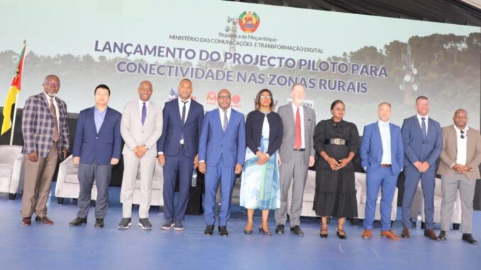 Lançamento do Projeto Piloto para Expansão da Telefonia Móvel nas Zonas Rurais de Moçambique Grupo de doze pessoas, entre homens e mulheres, em traje formal posando para foto oficial em evento do Ministério das Comunicações e Transformação Digital de Moçambique, com a bandeira de Moçambique ao fundo e um banner indicando o lançamento de projeto piloto para conectividade nas zonas rurais.