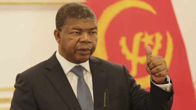 Presidente João Lourenço com terno escuro e gravata azul faz gesto de polegar para cima em frente à bandeira de Angola em um ambiente oficial.