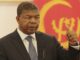 Presidente João Lourenço com terno escuro e gravata azul faz gesto de polegar para cima em frente à bandeira de Angola em um ambiente oficial.