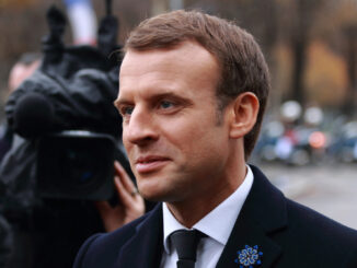 Emmanuel Macron em cerimônia oficial em Paris, usando terno azul, com bandeiras francesas ao fundo