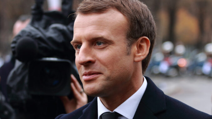 Emmanuel Macron em cerimônia oficial em Paris, usando terno azul, com bandeiras francesas ao fundo