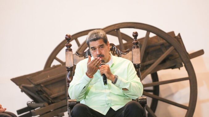 GPjb7b3XIAA34Fe Nicolás Maduro sentado em uma cadeira de madeira, segurando um microfone, gesticulando com a mão direita; ao fundo, uma carroça de madeira antiga.