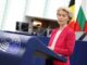 Presidente da Comissão Europeia Ursula von der Leyen vestindo blazer rosa e blusa branca, falando em um púlpito azul com o logotipo do Parlamento Europeu, com bandeiras nacionais ao fundo.