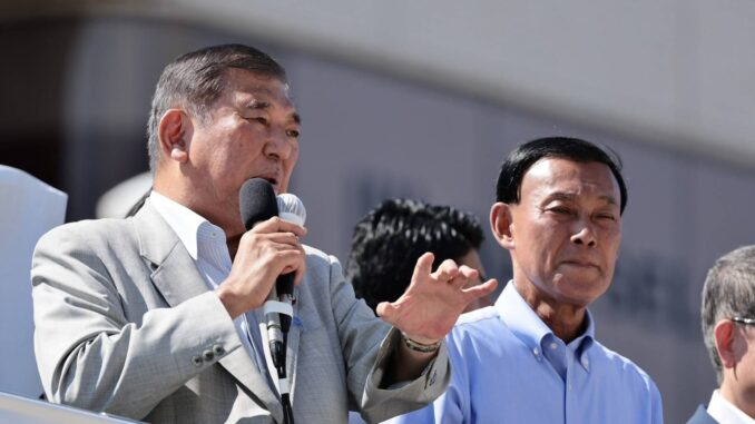 Gv0KGWtXUAMM1Rx Dois homens japoneses, um deles falando ao microfone durante um pronunciamento público.