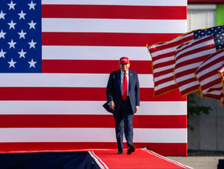 Donald Trump caminhando em um palco com bandeiras dos Estados Unidos ao fundo.