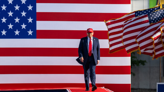 GvBKF4UXcAAKssd Donald Trump caminhando em um palco com bandeiras dos Estados Unidos ao fundo.