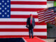 Donald Trump caminhando em um palco com bandeiras dos Estados Unidos ao fundo.