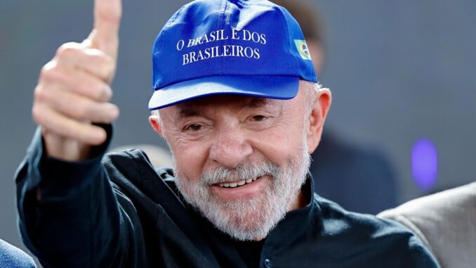O presidente Luiz Inácio Lula da Silva durante evento público Homem sorridente com barba branca usando boné azul escrito “O Brasil é dos brasileiros” e fazendo sinal de positivo com o polegar.