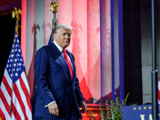 Donald Trump diante de colunas iluminadas em vermelho e azul, com bandeiras dos Estados Unidos ao fundo