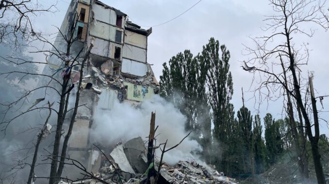 Escombros de Kiev: o dia em que a guerra bateu à porta Prédio parcialmente destruído em Kiev após ataque aéreo russo, com fumaça subindo dos escombros e árvores danificadas ao redor.