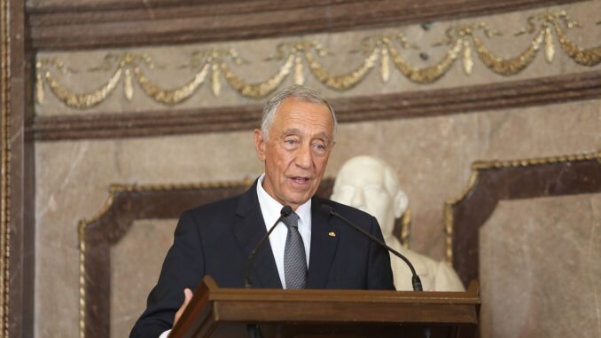 Marcelo Rebelo de Sousa em discurso oficial Presidente Marcelo Rebelo de Sousa discursando em um púlpito, em ambiente oficial, com estátua ao fundo.