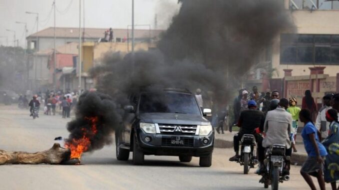 Carro preto atravessando rua com pneus em chamas e fumaça preta densa durante protestos em Luanda, com pessoas observando e motociclistas passando ao lado.