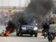 Carro preto atravessando rua com pneus em chamas e fumaça preta densa durante protestos em Luanda, com pessoas observando e motociclistas passando ao lado.