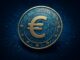 Imagem de uma moeda digital com o símbolo do euro (€) cercada por estrelas, sobre um fundo azul com padrões de circuito eletrônico, representando o euro digital e a tecnologia financeira na União Europeia.