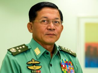 Retrato do General Min Aung Hlaing em uniforme militar verde com medalhas e insígnias, usando óculos e olhando diretamente para a câmera.