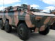 Veículo blindado EE-11 Urutu com camuflagem verde e marrom estacionado em área externa, sob céu azul com nuvens.