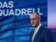 Friedrich Merz falando em um programa de televisão com fundo azul e o texto "Das Quadrell" ao fundo, usando terno azul e gravata clara.