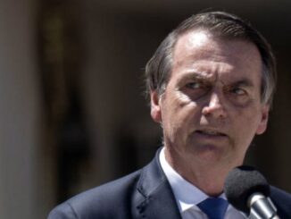 Jair Bolsonaro discursando em microfone, terno azul, expressão séria, em evento ao ar livre.