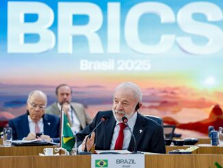 Presidente Lula discursando em reunião do BRICS no Brasil em 2025, com a bandeira brasileira à frente e painel com o logo do BRICS ao fundo.
