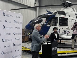 Senador Roger Wicker falando em um púlpito com logotipo da UMMC, com um helicóptero AirCare ao fundo e fotógrafos registrando o evento.
