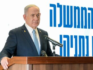 Benjamin Netanyahu em discurso oficial, vestindo terno escuro e gravata azul clara, falando em um púlpito com microfones, com fundo branco e texto em hebraico azul.