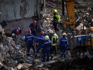 Equipe de resgate e bombeiros em uniformes azuis e capacetes vermelhos e amarelos trabalhando entre destroços e escombros de um prédio destruído em Kiev após ataque aéreo.