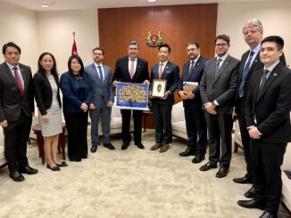 Foto de uma delegação empresarial brasileira em Singapura. Eduardo Riedel (PSDB) está ao centro, segurando uma obra de arte, rodeado por nove representantes de Singapura e empresários. Todos vestem trajes formais e posam em um ambiente institucional, com bandeira e brasão ao fundo.