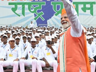 Primeiro-ministro Narendra Modi acena para a multidão durante celebração do Dia da Independência da Índia, com jovens participantes vestidos de branco ao fundo e banner “Nation First”.