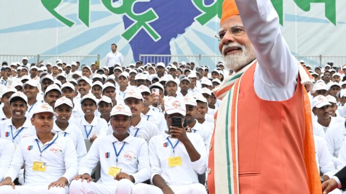 Dia da Independência 2025: Modi reforça unidade e segurança nacional Primeiro-ministro Narendra Modi acena para a multidão durante celebração do Dia da Independência da Índia, com jovens participantes vestidos de branco ao fundo e banner “Nation First”.