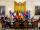 Líderes internacionais, incluindo Donald Trump e Volodymyr Zelensky, em reunião na Sala Leste da Casa Branca com bandeiras de EUA, Ucrânia, Alemanha, França, Finlândia, Itália, Reino Unido, OTAN e União Europeia ao fundo.