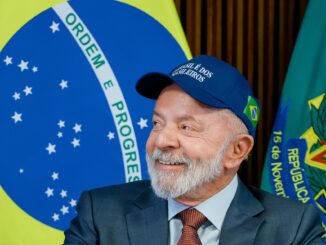 Presidente Luiz Inácio Lula da Silva usando boné azul com a frase "O Brasil é dos brasileiros", sorrindo, com a bandeira do Brasil ao fundo.
