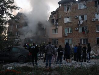 Pessoas observam bombeiros trabalhando enquanto fumaça sai de um prédio residencial danificado em Kiev após ataque com mísseis russos.