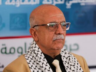 Foto do primeiro-ministro houthi Ahmed Ghaleb al-Rahawi usando óculos, gravata e lenço palestino, em evento oficial em Sanaa, Iémen.