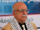 Foto do primeiro-ministro houthi Ahmed Ghaleb al-Rahawi usando óculos, gravata e lenço palestino, em evento oficial em Sanaa, Iémen.