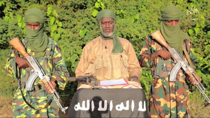 Ameaça Extremista na África: Expansão e Impactos dos Grupos Jihadistas no Sahel e Leste Africano Três combatentes armados com rifles automáticos, dois deles com rostos parcialmente cobertos por lenços verdes, posam ao redor de um homem central que está sentado atrás de uma mesa coberta por um pano preto com inscrições em árabe, em ambiente externo com vegetação ao fundo.